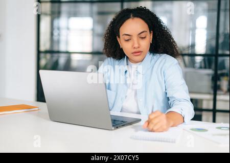 Konzentrierte, kluge, beschäftigte brasilianische oder hispanische lockige Frau in einem pastellblauen Hemd, Manager, Sekretärin, sitzt in einem modernen Büro am Schreibtisch, arbeitet an einem Laptop, macht Notizen im Notebook, plant Stockfoto