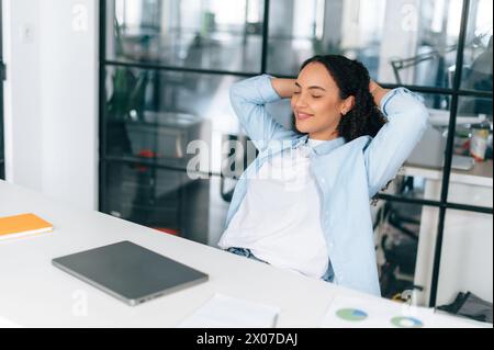 Ruhen Sie sich während des Arbeitstages aus. Glückliche brasilianische oder hispanische Frau, Mitarbeiterin des Unternehmens, entspannte sich am Arbeitsplatz, beendete die Arbeit am Laptop im Büro, legte ihre Hände hinter den Kopf und schloss lächelnd die Augen Stockfoto