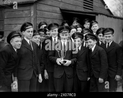 FIFTH SEA LORD VERLEIHT AUSZUBILDENDEN AUS DER FLOTTE PREISE. 15. UND 16. DEZEMBER 1943, ROYAL MARINEFLUGSTRAININGS, NEWCASTLE-UNDER-LYME. DER FÜNFTE SEA LORD UND CHIEF OF MARINE AIR EQUIPMENT, KONTERADMIRAL D W BOYD, CBE, DSC, ÜBERREICHTE PREISE FÜR DIE AUSSCHEIDUNGSZEIT DER FLOTTENLUFTWAFFENAUSZUBILDENDEN BEI DER R N A T E - THE BEST LOSER; APPRENTICE P E TUCKER OF SHOEBURYNESS; vor seiner Hütte mit einigen seiner Kameraden. Er ist stolz darauf, die Trophäe „Bester Verlierer“ des Fünften Meeres zu erhalten, die jedes Jahr im Establishment ausgetragen wird Stockfoto