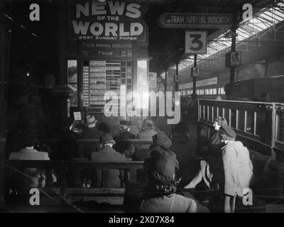 BRITISH RAILWAYS AT WAR - HALLO UND AUF WIEDERSEHEN: TRANSPORT AND TRAVEL IN WARTIME, UK, 1944 - Männer und Frauen warten darauf, Freunde und Verwandte aus einem Zug zu treffen, sitzen in der Nähe des Ankunftsindikators neben Bahnsteig 3, bis die entsprechende Bahnsteignummer angezeigt wird. Unter einer Werbung für die Zeitung „News of the World“ sind die Orte, von denen Züge ankommen, und die geplanten Ankunftszeiten auf der Anzeigetafel aufgeführt. Der Zug von Liverpool über Crewe ist beispielsweise um 14:20 Uhr fällig, der Zug von Blackpool über Preston, Wigan und Crewe um 40 Uhr. Eine Uhr ist nur für den sichtbar Stockfoto