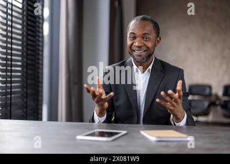 Ein Mann im Anzug sitzt mit einem Tablet und einem Notizbuch an einem Schreibtisch. Er lächelt und führt ein Gespräch Stockfoto