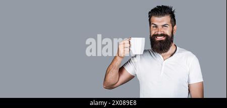 Lächelnder bärtiger Hipster-Mann, der eine Tasse Kaffee oder Tee hält. Gutaussehende Männer halten eine Tasse Kaffee und Tee. Bärtiger Mann lächelt. Guten Morgen, Mann Tee. Lächelnd Stockfoto