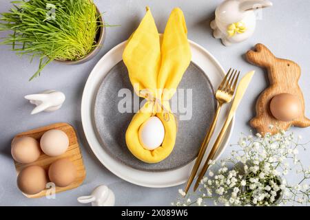 Ei in gelber Serviette als Hasenohren auf Tellern, Eierständer aus Holz mit Bio-Eiern, sanfte weiße Blüten, frisches Gras, Keramikhasen, Goldbesteck drauf Stockfoto