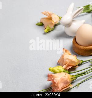 Trendige Bio-Osterkarte. Natürliches beiges Ei im hölzernen Eierständer, sanfte Pfirsichblüten Eustoma, weißer Keramikhase auf grauem Hintergrund. Noch Urlaub Stockfoto