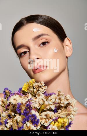 Eine brünette junge Frau strahlt Schönheit aus, während sie einen Blumenstrauß in einem Atelier vor grauem Hintergrund zärtlich hält. Stockfoto