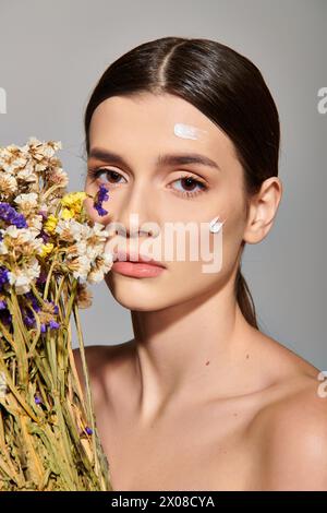 Eine junge Frau mit brünetten Haaren hält einen blühenden Blumenstrauß, der Schönheit und Eleganz in einem Studio vor grauem Hintergrund versprüht. Stockfoto