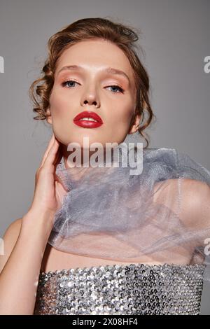 Eine junge Frau, die klassische Schönheit in einem silbernen Kleid und einem auffälligen roten Lippenstift ausstrahlt und selbstbewusst in einem Studio vor grauem Hintergrund posiert. Stockfoto