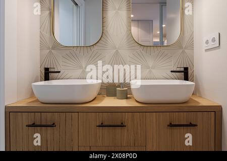 Ein moderner Kosmetikbereich im Bad mit zwei ovalen Waschbecken, mattschwarzen Armaturen, einer strukturierten Wand mit Akzenten und einem eleganten Spiegel mit goldenem Rahmen Stockfoto