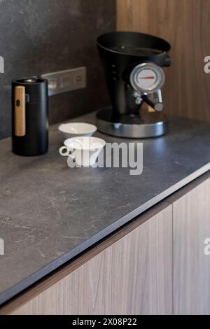 Die moderne Küchenecke verfügt über eine elegante schwarze Espressomaschine, ein Mahlwerk und Keramikbecher auf einer dunklen Arbeitsfläche Stockfoto