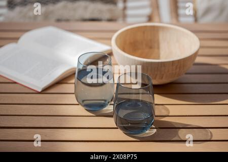 Eine gemütliche Sommerszene mit einem offenen Buch und zwei stiellosen Weingläsern gefüllt mit Wasser auf einem hölzernen Terrassentisch, begleitet von einem leeren Holzbogen Stockfoto