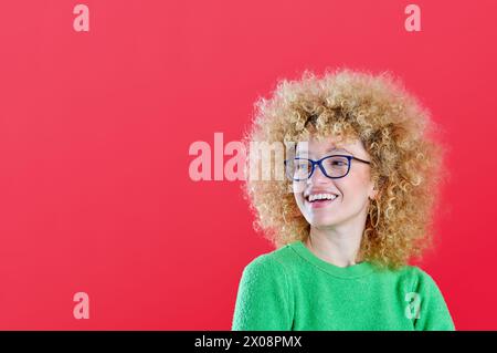 Eine fröhliche Frau mit flauschigem lockigem Haar und Brille lächelt zur Seite und trägt einen grünen Pullover vor einem kontrastierenden roten Hintergrund Stockfoto