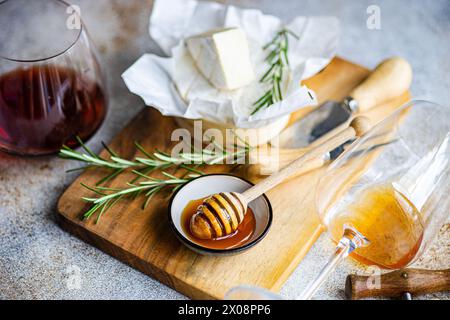 Eine Gourmet-Auswahl mit Brie-Käse, frischem Rosmarin, Honigdipper und Gläsern Rot- und Weißwein, elegant vor einem strukturierten Hintergrund Stockfoto