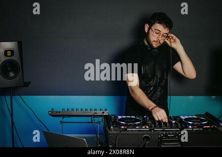 Junger männlicher DJ mit Kopfhörern um den Hals konzentriert, während er die Einstellungen an einem Mixer in einer dunklen Club-Umgebung anpasst Stockfoto