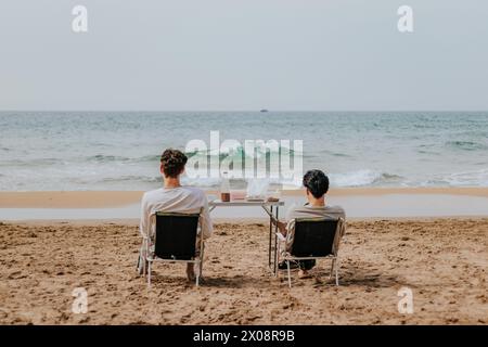 Rückansicht von zwei Personen, die auf Stühlen mit Blick auf das Meer sitzen und eine ruhige Strandlandschaft mit einer Weinflasche und Gläsern auf einem Tisch zwischen ihnen genießen. Stockfoto