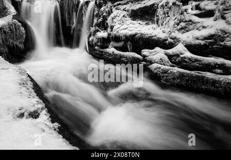 Ein ruhiges Schwarz-weiß-Foto, das einen Fluss einfängt, der durch eine schneebedeckte Berglandschaft fließt, mit einem kaskadierenden Wasserfall und eisigen Felsen. Stockfoto