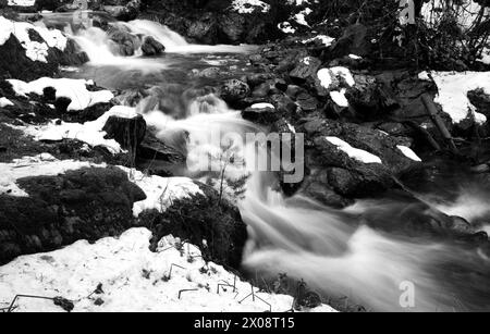 Ein Schwarzweiß-Foto, das den dynamischen Fluss eines Flusses einfängt, der durch eine schneebedeckte Hochgebirgslandschaft schneidet. Stockfoto