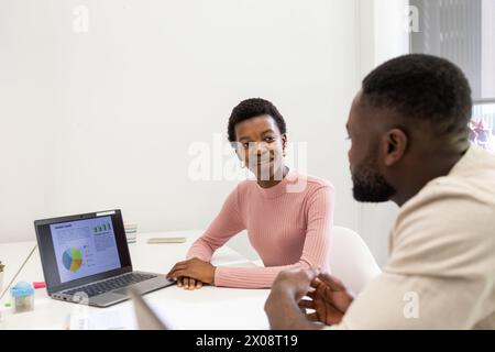 Afroamerikanerin lächelt während eines Meetings mit einem männlichen Kollegen in die Kamera, mit einem Laptop, der Karten auf dem Tisch zeigt. Stockfoto