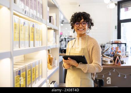 In ihrer Boutique steht eine fröhliche Eigentümerin eines kleinen Unternehmens und hält ein digitales Tablet in den Regalen mit Produkten. Stockfoto