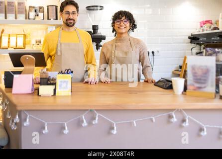 Zwei fröhliche Kaffee-Multiethnische Angestellte in Schürzen stehen hinter der Theke in einem gemütlichen Café. Stockfoto