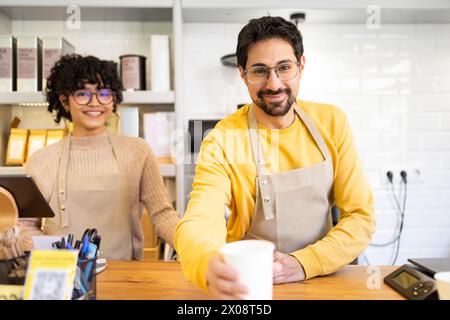 Zwei fröhliche multiethnische Baristas mit warmem Lächeln stehen hinter der Theke in einem modernen Café und bieten eine frisch gebrühte Tasse Kaffee an. Stockfoto