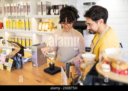 Zwei lächelnde multiethnische Barista bereiten gemeinsam Kaffee in einem modernen Kaffee zu, umgeben von Kaffeeprodukten und -Geräten. Stockfoto