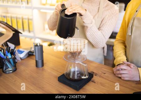 Ein Barista mit einem schwarzen Wasserkocher gießt heißes Wasser über Kaffeesatz in einem Tropfer aus Glas, während ein Gast wartet. Stockfoto