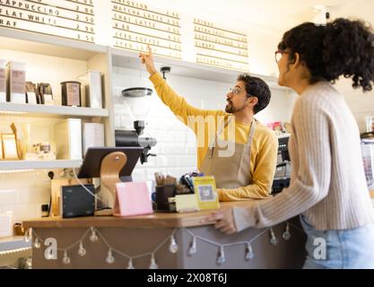 Der Barista auf der Schürze zeigt dem Gast in einem gemütlichen Café auf das Menü. Stockfoto