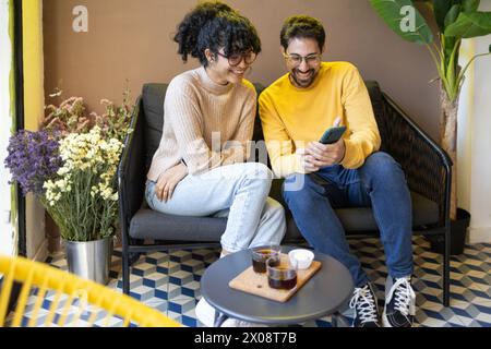 Ein fröhliches, multiethnisches Paar sitzt eng auf einer Couch, vertieft in einem Smartphone-Bildschirm, lacht in einer gemütlichen Innenumgebung mit dekorativen Blumen nea Stockfoto