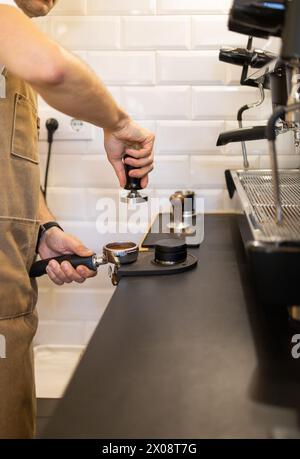 Ein Barista in einer Schürze drückt gemahlenen Kaffee in einen Portafilter neben einer Espressomaschine und zeigt eine Nahaufnahme der Hände und Werkzeuge, die im Kaffee verwendet werden Stockfoto