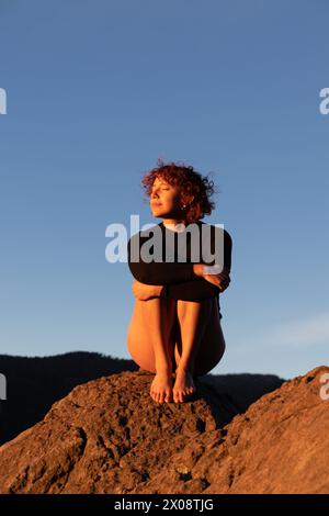 Eine moderne Tänzerin sitzt nachdenklich auf einem zerklüfteten Felsen, getaucht in das weiche, warme Licht der untergehenden Sonne, vor einem klaren blauen Himmel Stockfoto