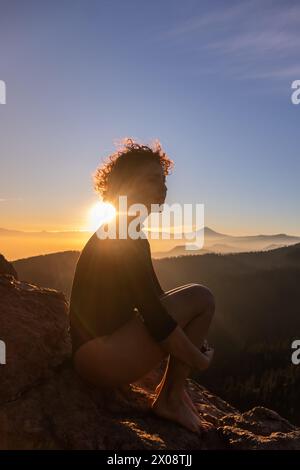 Eine Person mit lockigen Haaren hat eine Silhouette vor dem Sonnenuntergang hinter den Bergen, was eine ruhige und ruhige Szene schafft Stockfoto
