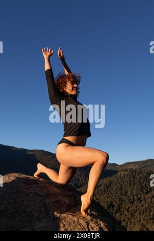 Eine junge Tänzerin posiert in der Abenddämmerung auf einem Berg und zeigt Freiheit und Ausdruck unter dem klaren blauen Himmel Stockfoto