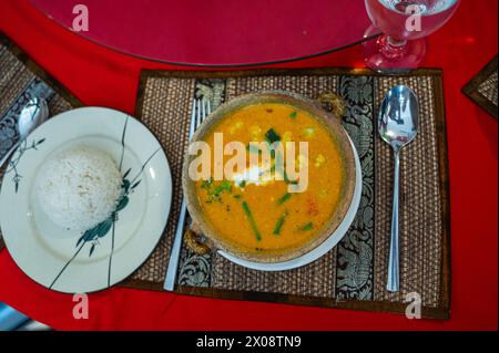 Von oben traditionelle kambodschanische Suppe, serviert in einer rustikalen Schüssel mit einer Seite Reis, auf einem gewebten Tischset und einer roten Tischdecke. Stockfoto