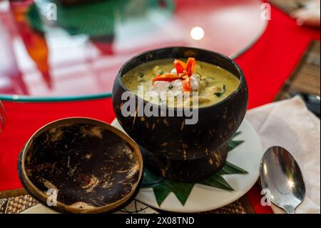Eine duftende kambodschanische Suppe in einer natürlichen Kokosnussschale, garniert mit einer roten Chili-Blume, auf einer roten Tischplatte. Stockfoto