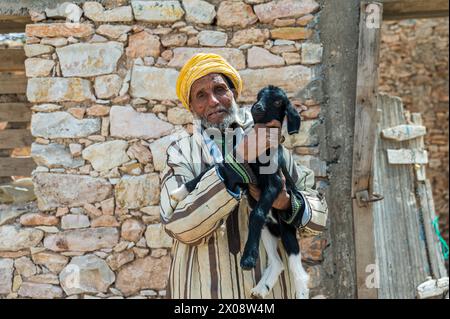 Ein älterer marokkanischer Mann, der einen traditionellen Turban trägt, hält eine kleine schwarze Ziege im Arm vor dem Hintergrund einer rustikalen Steinmauer Stockfoto