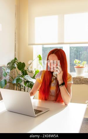 Eine lächelnde rothaarige Frau, die freudig am Telefon einen Ausflug in die Natur plant, mit offenem Laptop vor ihr in einem hellen, sonnendurchfluteten Raum Stockfoto