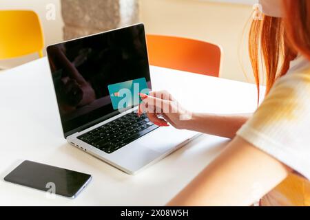Anonyme rothaarige Frau ist mit Online-Shopping beschäftigt, hält eine Kreditkarte in der Nähe eines Laptops, während sie an einem modernen Tisch sitzt und hebt E-Commerce hervor Stockfoto