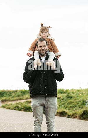 Ein fröhliches Kleinkind macht einen Huckepack-Ritt auf den Schultern ihres Vaters während eines entspannten Spaziergangs im Freien Stockfoto