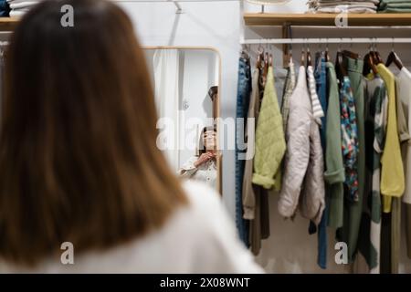 Eine Frau untersucht ihre Reflexion, während sie ein Frühlingsoutfit in einer Boutique-Umkleidekabine anprobiert, die von saisonalen Kleidungsstücken umgeben ist Stockfoto