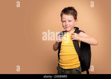 Ein lächelnder kleiner Junge mit Rucksack steht selbstbewusst vor beigefarbenem Hintergrund und ist bereit für einen Schultag Stockfoto