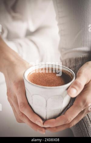 Eine Person, die einen Becher mit einer köstlichen und schaumigen heißen Schokolade in einer gemütlichen, warmen Umgebung hält, die das Wesen des Komforts einfängt Stockfoto