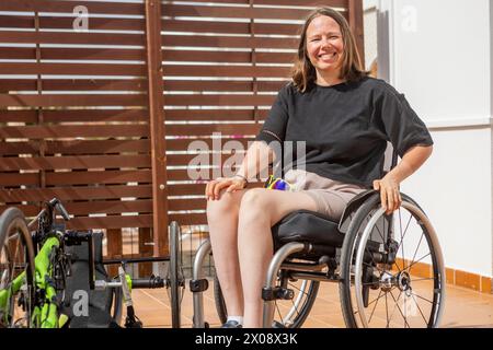 Fröhliche behinderte Frau sitzt im Rollstuhl neben ihrem adaptiven Handrad und zeigt Entschlossenheit und Leichtathletik. Stockfoto