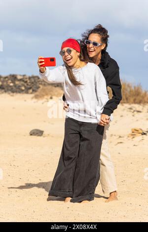 Ein fröhliches Paar lacht, während es ein Selfie an einem Sandstrand macht, um einen fröhlichen Moment ihres Campingausflugs festzuhalten Stockfoto