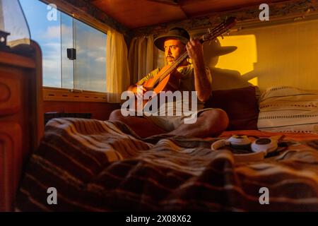 Ein nachdenklicher männlicher Musiker spielt eine Gitarre in seinem gemütlichen Wohnmobil, umgeben von warmem Sonnenuntergangslicht und inspiriert von den nahe gelegenen Meeresausblicken. Stockfoto