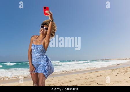 Eine Frau genießt es, ein luftiges Selfie an den Meereswellen zu machen, ihre Freude an einem perfekten, sonnigen Strandtag Stockfoto
