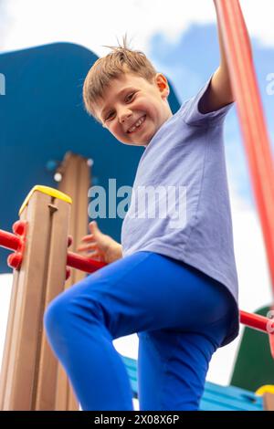 Von unten aus genießt ein fröhlicher kleiner Junge auf einem farbenfrohen Spielplatz die Spielzeit und klettert mit einem hellen Lächeln auf die Ausrüstung. Stockfoto