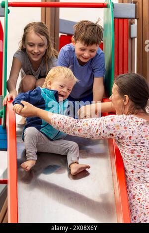 Eine lebendige und herzerwärmende Szene, während eine Familie die Zeit zusammen auf einer Spielplatzrutsche genießt. Die pflegende Mutter hilft dem Kind, das begeistert ist, und zeigt eine Funktion Stockfoto