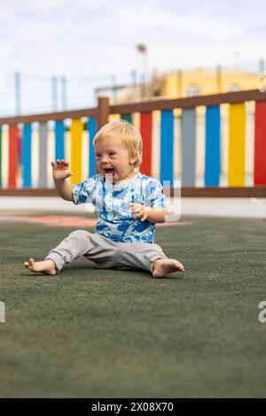 Fröhlicher Junge mit Down-Syndrom, der auf der Spielplatzoberfläche sitzt, lacht und spielt allein, mit buntem Zaunhintergrund. Stockfoto