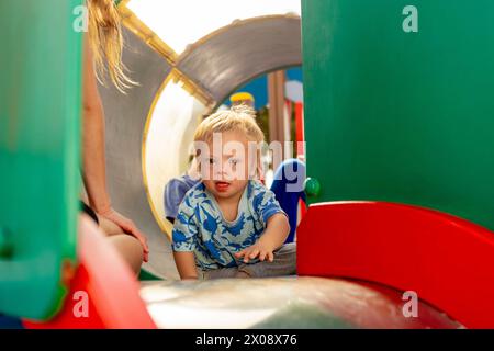 Ein kleiner Junge mit Down-Syndrom genießt Spielzeit in einem Spielplatztunnel mit Familienmitgliedern und verkörpert Inklusion und familiäre Liebe. Stockfoto