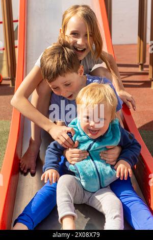 Glückliche Geschwister spielen auf einer Folie mit ihrem jüngeren Bruder, der an Down-Syndrom leidet, und zeigen einen fröhlichen, inklusiven Familienmoment im Freien. Stockfoto